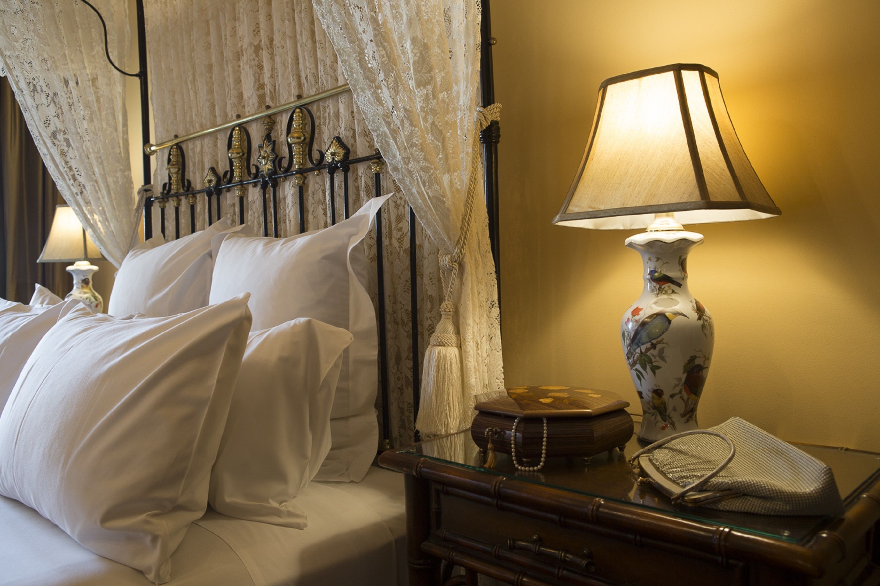 A neatly made bed with white linens, a lace canopy, and a bedside lamp with a floral vase.