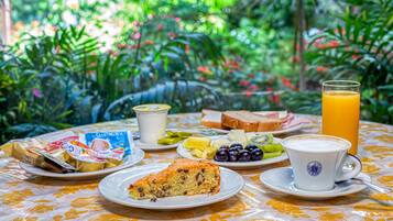 Una mesa de desayuno con diversos alimentos, como tostadas, fruta y una rebanada de pastel, con un fondo de exuberante follaje.