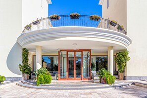 A hotel entrance with a curved balcony, glass doors, and potted plants. - Hotel Le Torri (Arborea)