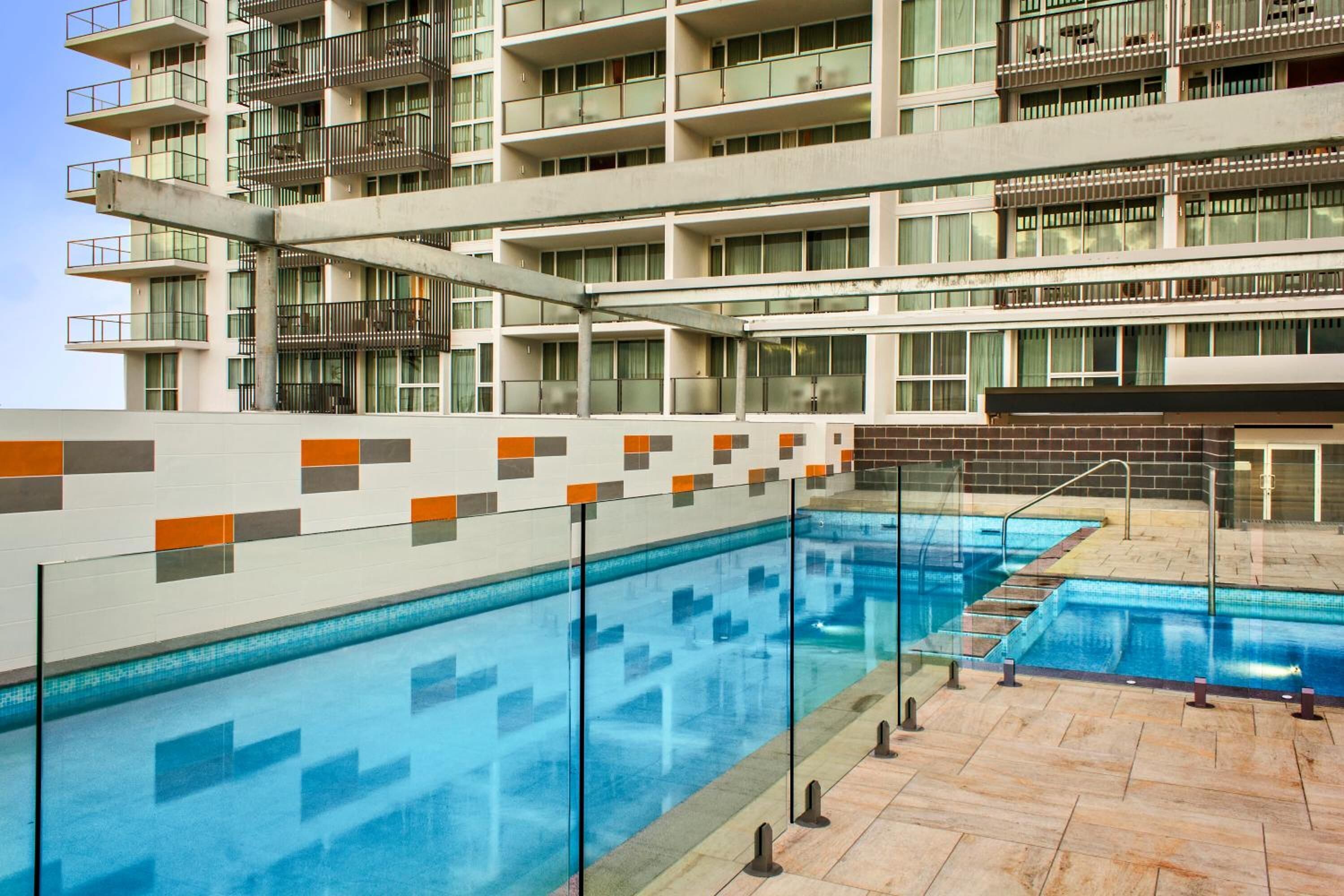 Un hotel de varios pisos con zona de piscina, con un diseño moderno que incluye barandillas de vidrio y un camino empedrado.