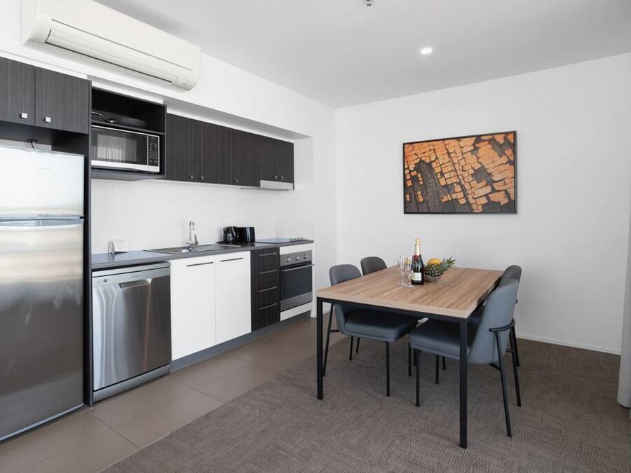 A modern kitchen with a built-in microwave, stainless steel refrigerator, and dishwasher. There is a dining table with chairs and a wooden floor.