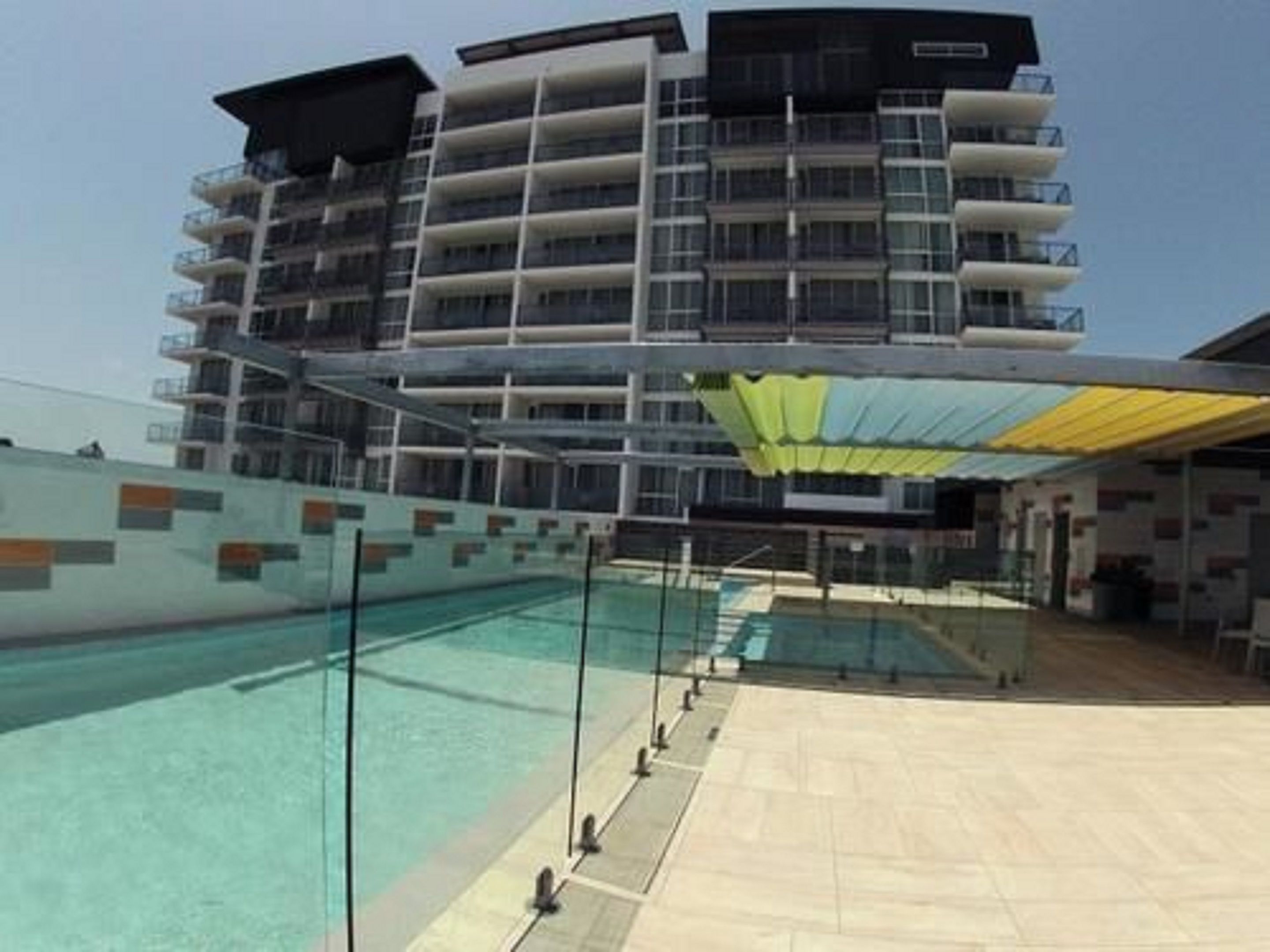 Un hotel moderno con una amplia piscina exterior, varios balcones y una zona cubierta con un toldo amarillo.