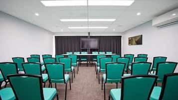 Uma sala de conferências com fileiras de cadeiras verdes, uma TV de tela plana grande e piso coberto de carpete.