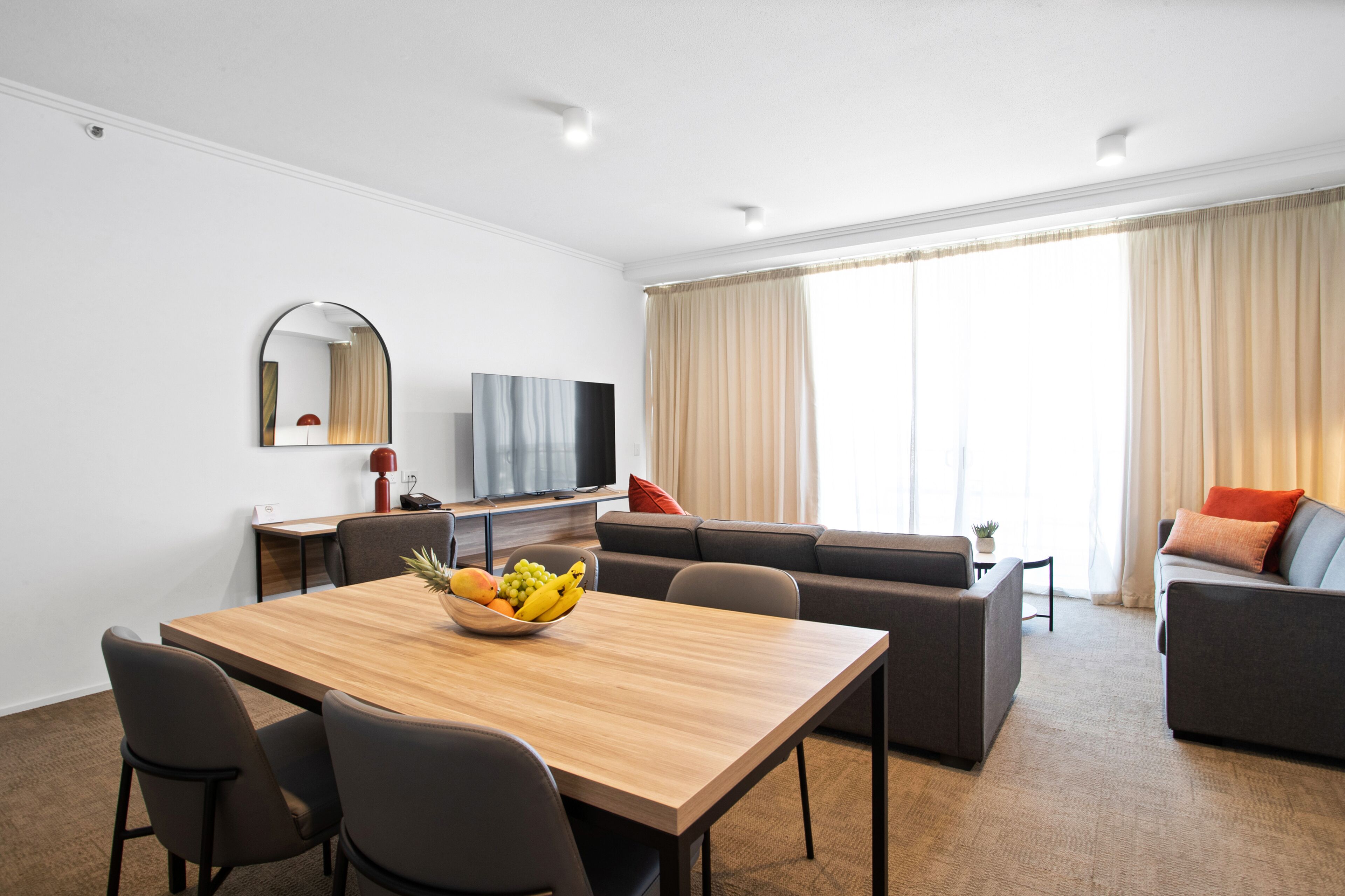 A modern living room with a wooden dining table, grey sofas, a flat-screen TV, and a large window with curtains.