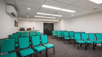 A conference room with rows of teal chairs, a carpeted floor, a white ceiling with fluorescent lights, and a door with an emergency exit sign.