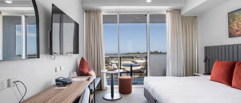 Habitación de hotel con cama, escritorio, silla, televisor, balcón con vistas y ventana con cortinas.