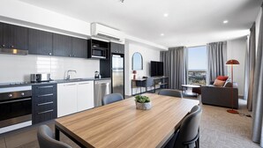 A modern kitchen with dark cabinets, a wooden dining table, and a city view through large windows. - Quest Mackay on Gordon (Mackay)