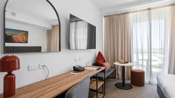 A hotel room with a wooden desk, a large mirror, a flat-screen TV, a bed, a small round table with fruit, and a window with curtains.