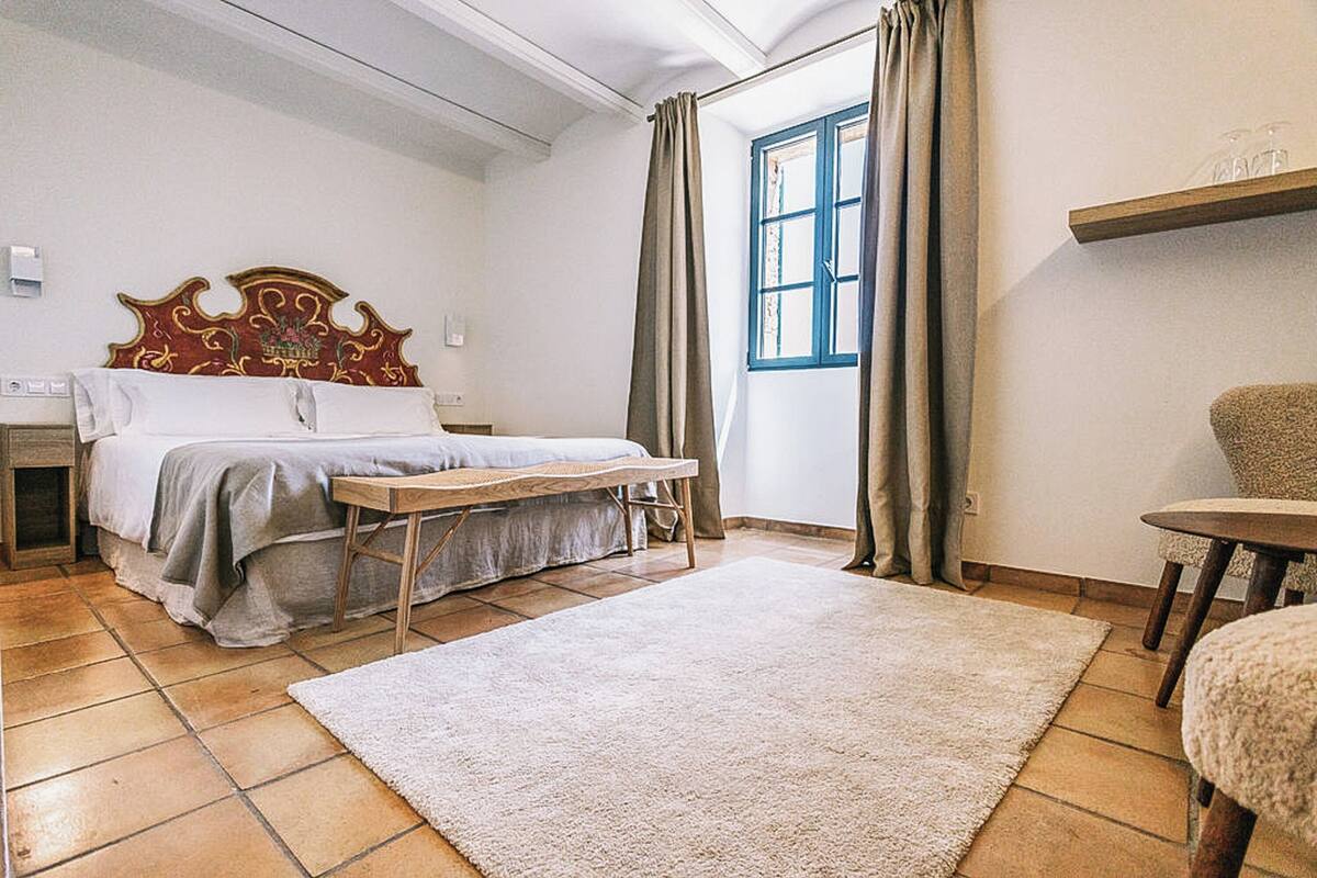 Quarto com cama, mesa de cabeceira, cadeira, janela com cortinas e cabeceira de madeira.