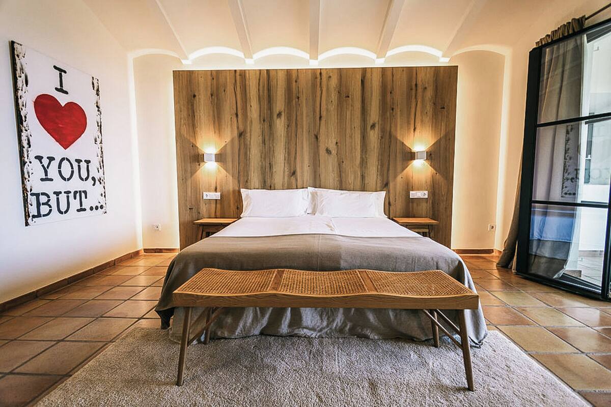 Quarto com cabeceira de madeira, duas luminárias de cabeceira, cama com roupa de cama branca, um banquinho de palha e uma janela grande com cortinas.