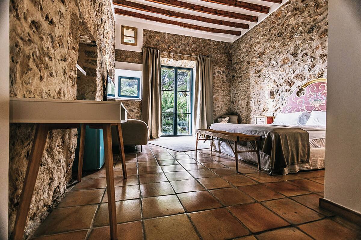 Una habitación rústica con paredes de piedra, una mesa de madera, una cama con cabecera rosa y vistas a la vegetación por una ventana.