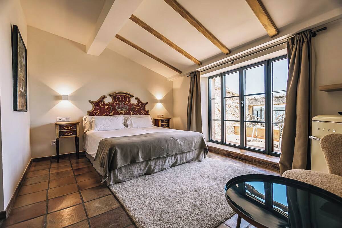 Quarto com cama grande, duas mesas de cabeceira, um criado-mudo, uma cadeira, janela com cortinas e vista para o exterior.