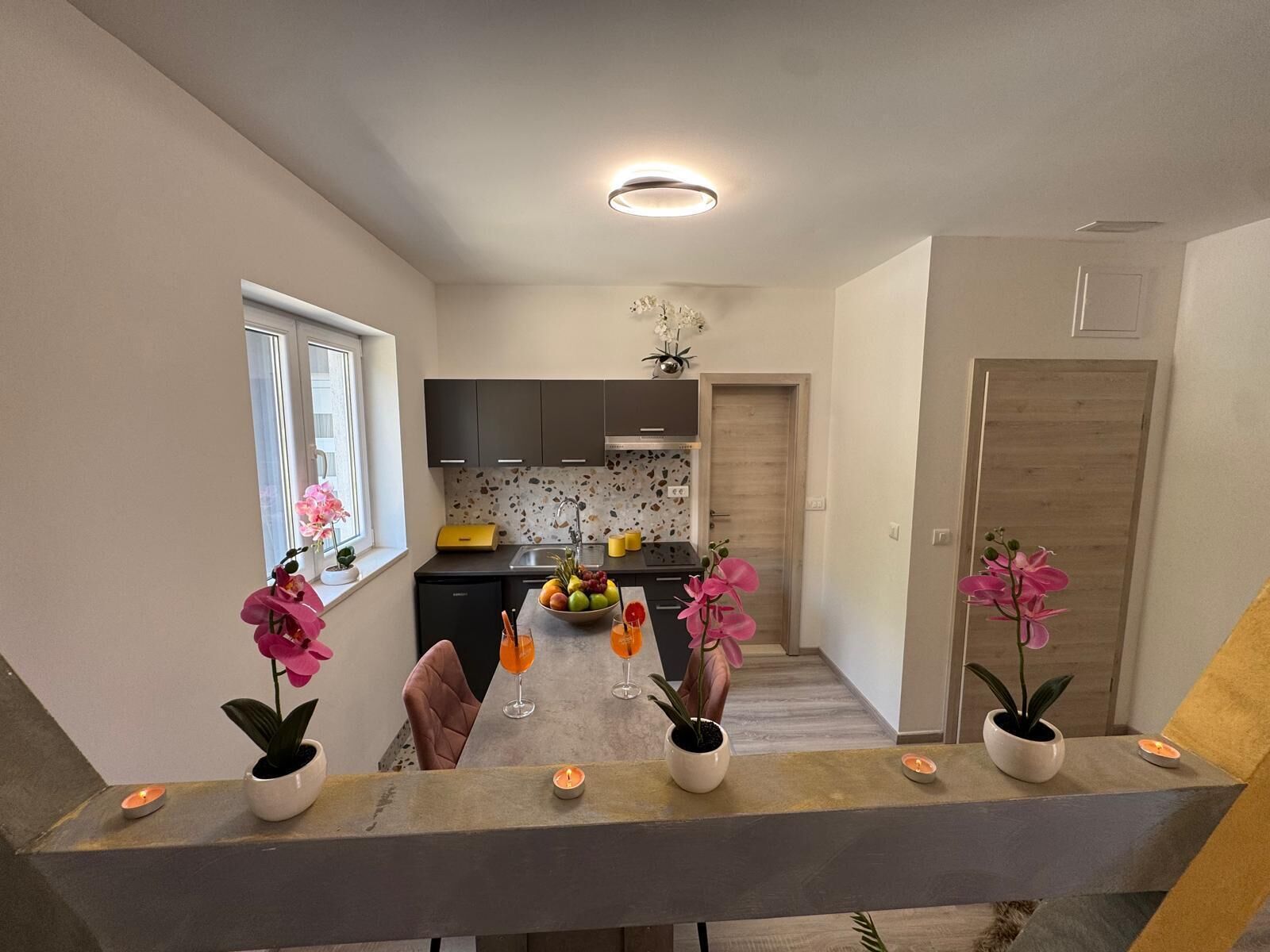 Une cuisine moderne avec des meubles de couleur sombre, un plan de travail et un revêtement mural. On peut y voir des plantes en pot et des bougies sur la table à manger.