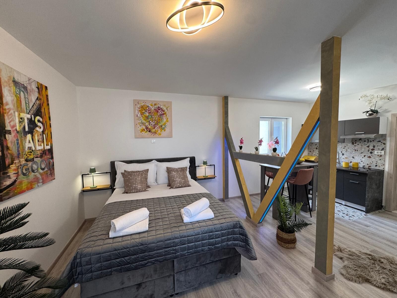 A modern bedroom with a bed, bedside tables, a painting, and a staircase leading to a kitchen area.