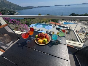 Un balcon avec une table préparée pour prendre un verre et déguster des fruits, donnant sur une piscine et offrant une vue panoramique sur la mer.
