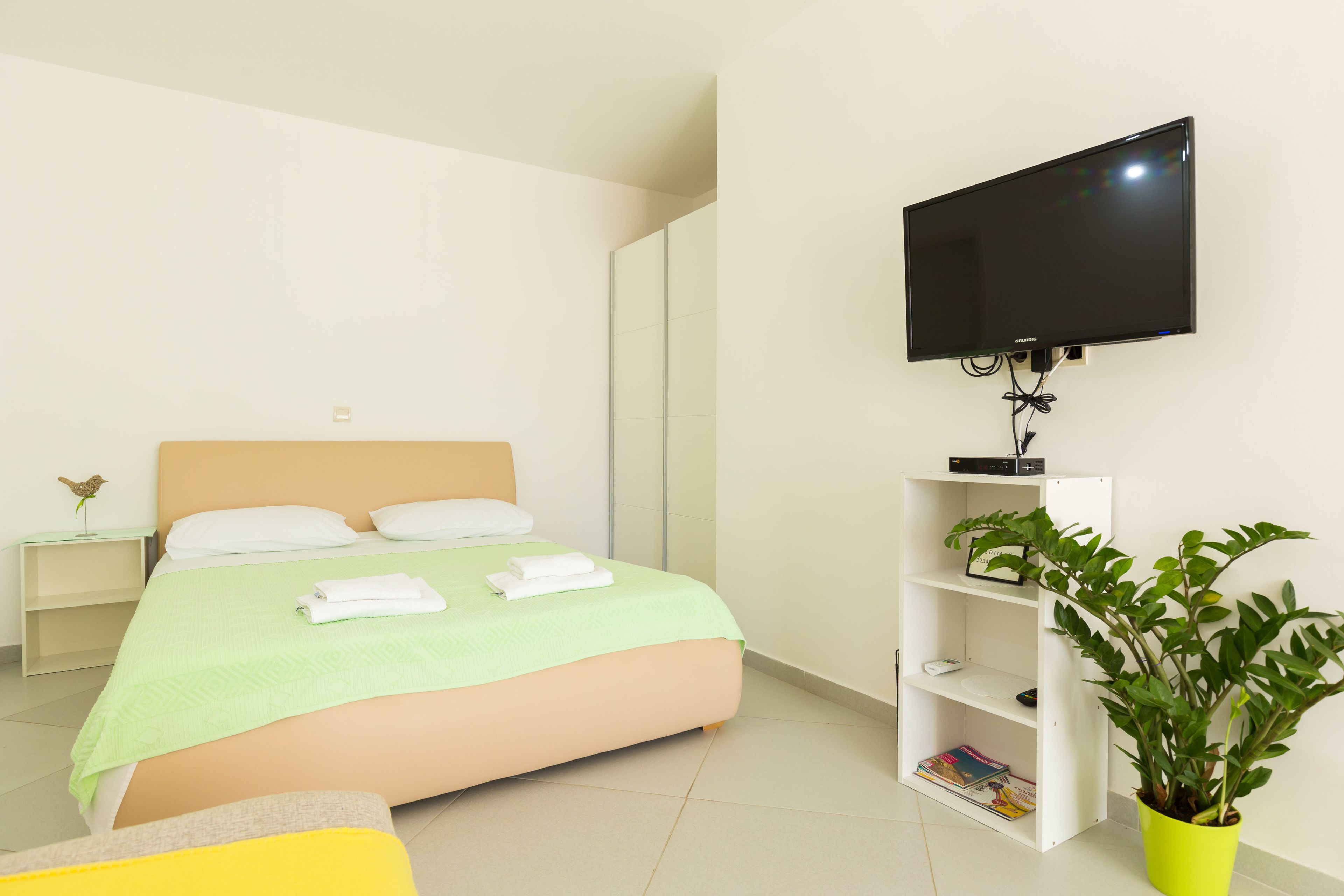 Quarto de hotel com uma cama, televisão na parede, mesa de cabeceira com abajur e planta em vaso.