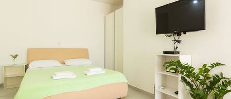 Quarto de hotel com uma cama, televisão na parede, mesa de cabeceira com abajur e planta em vaso.