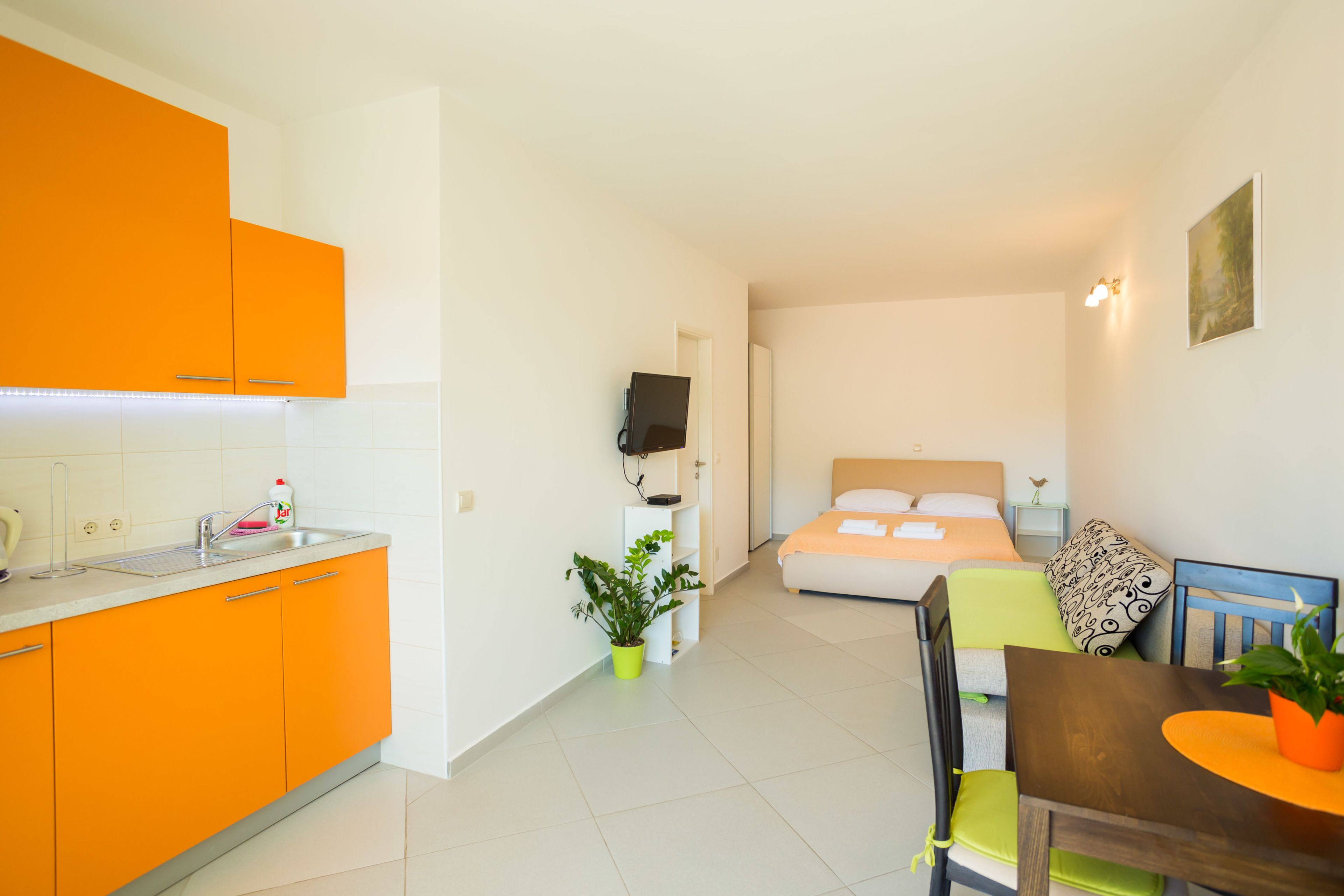A compact hotel room with a kitchenette, a bed with a headboard, a dining table with chairs, and a small potted plant.