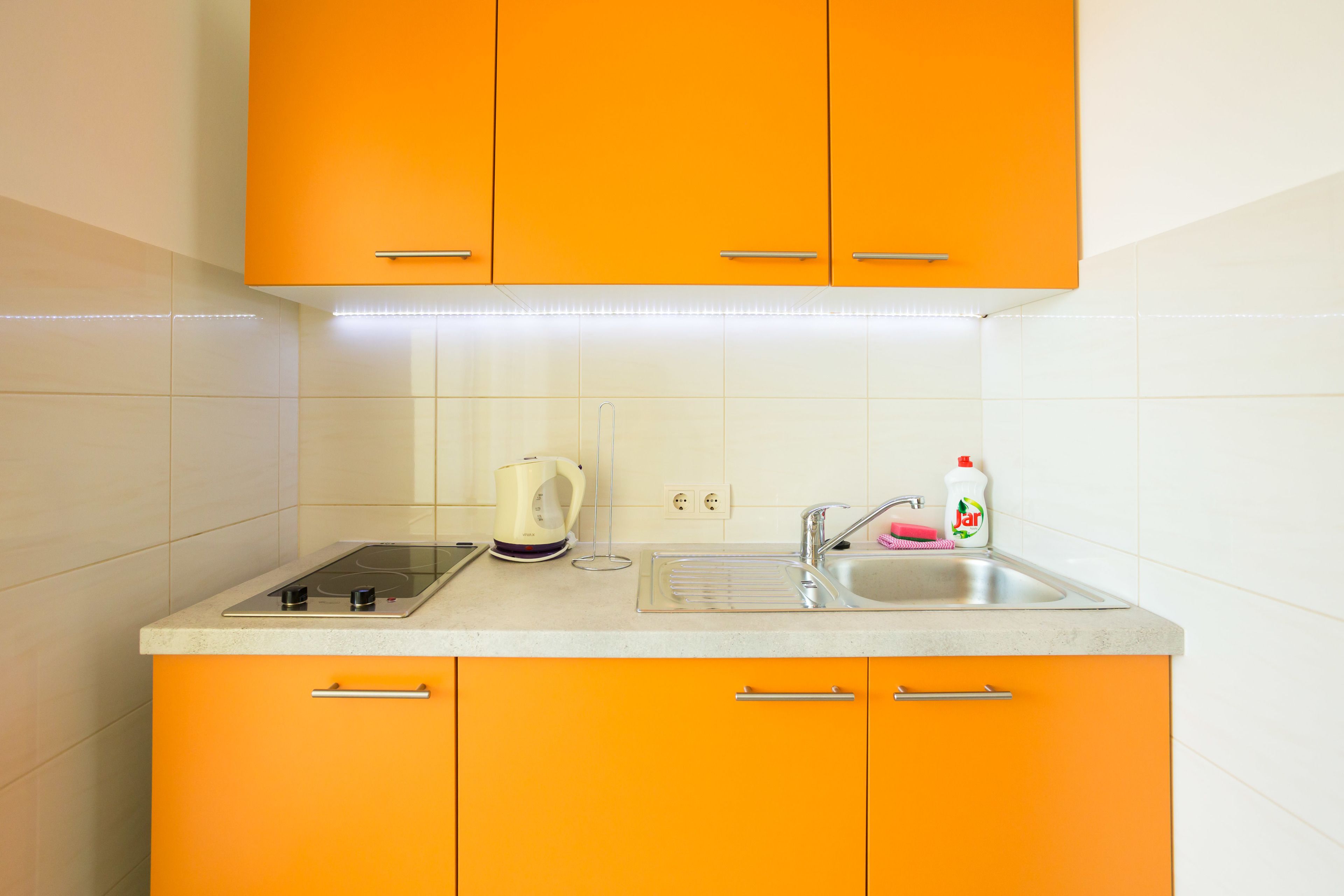 A kitchen with orange cabinets, a white countertop, a kettle, a sink, and a dish soap bottle.