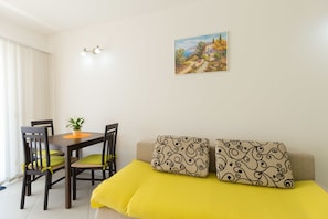 Um quarto com uma mesa de jantar, cadeiras, um sofá com almofadas estampadas, um quadro na parede e uma planta em vaso.