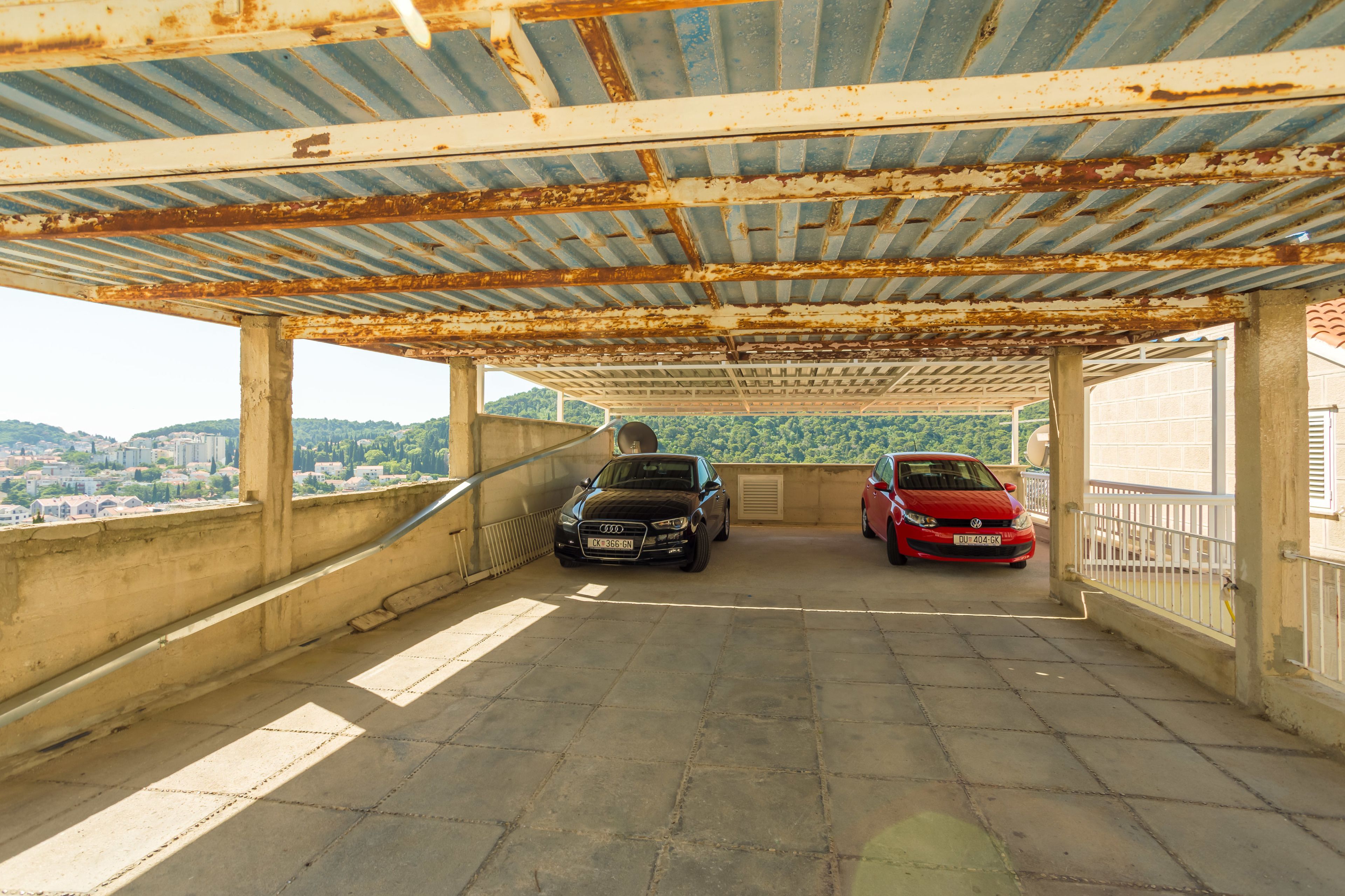 An outdoor covered parking area with two cars parked, a view of a cityscape in the background, and a rusty metal roof.