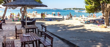 A beachside café with wooden tables and benches, overlooking a clear blue sea with people sunbathing and swimming.