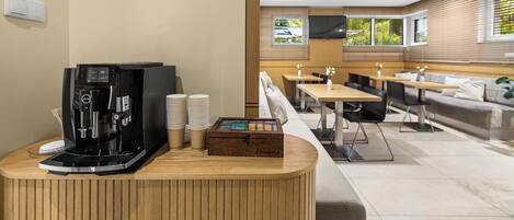 A modern cafe with a coffee machine, stacked cups, and a wooden box on a counter. There are tables and chairs in the background with a view of the outdoors.