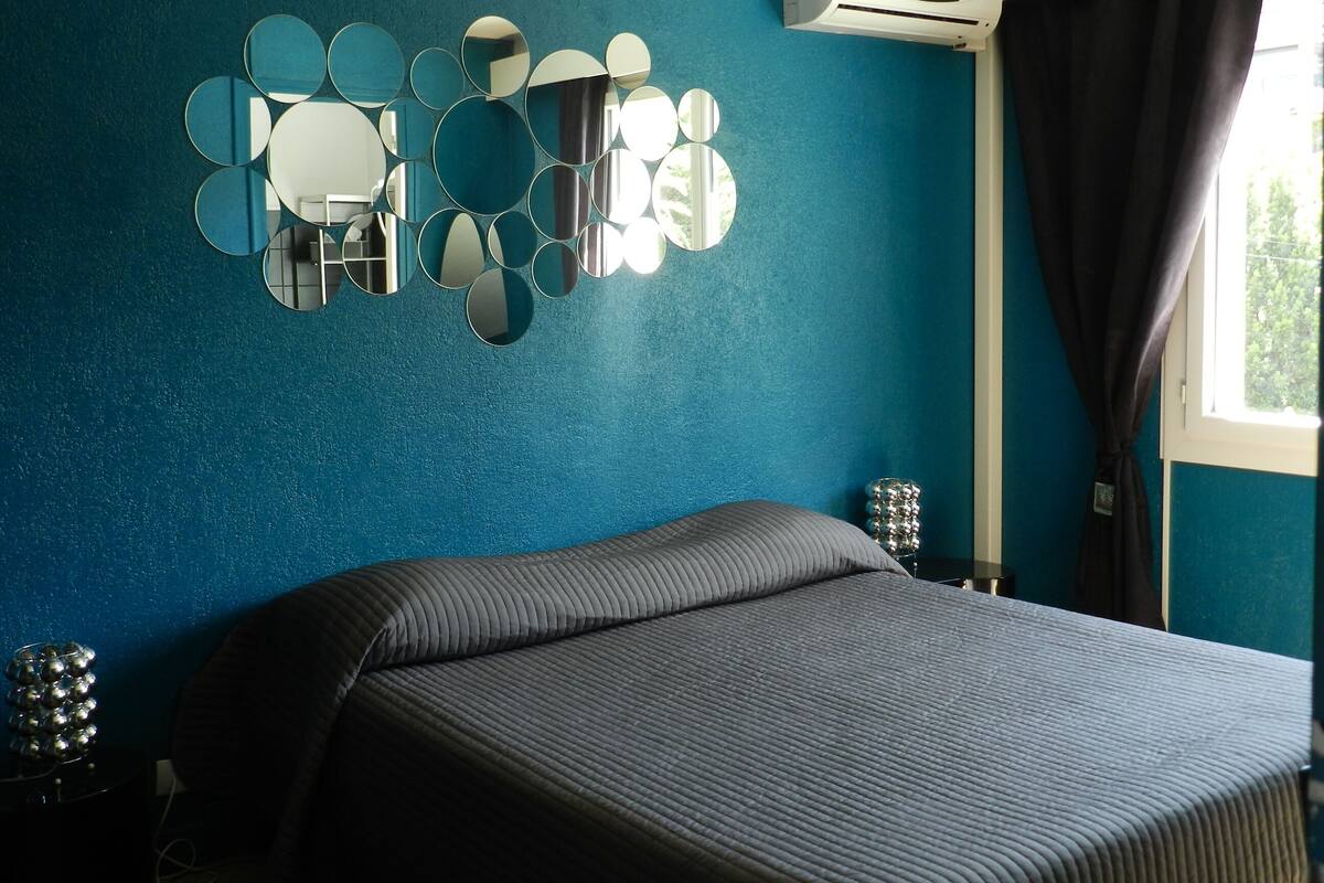 A bedroom with a bed, a wall-mounted mirror arrangement, an air conditioner, and a window with curtains.