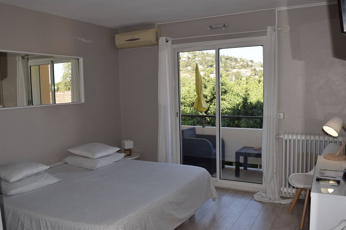 A bedroom with a bed, a small bedside table, a chair, a radiator, and a view of a balcony with a table and chairs.