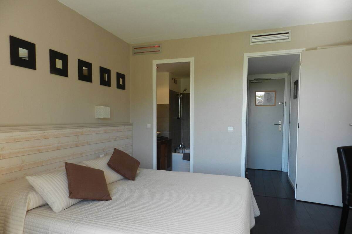 A hotel room with a large bed, a nightstand, a wall-mounted lamp, and a door leading to a bathroom.