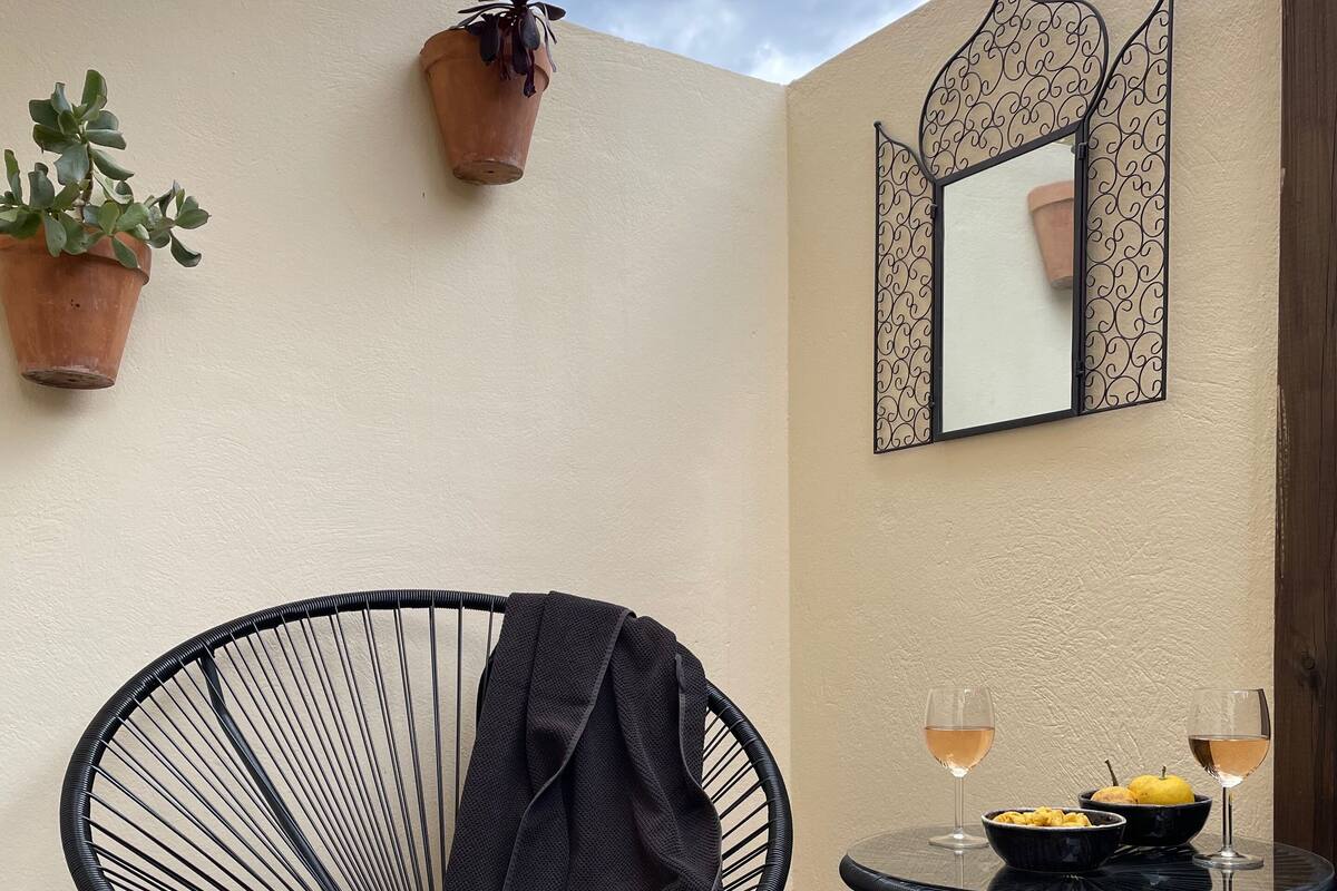 A black wire chair with a jacket draped over it, a small round table with a bowl of fruit and two glasses of wine, and a decorative mirror on the wall.