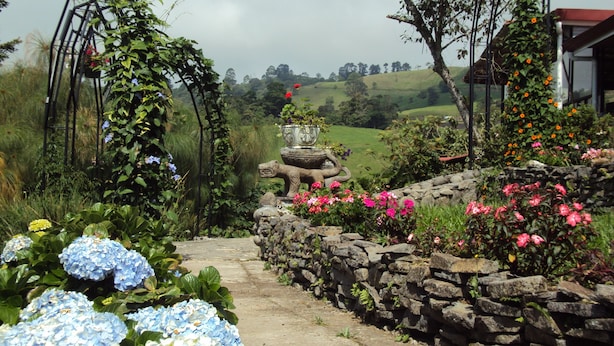Un sentier de jardin bordé de murs en pierre et agrémenté de différentes fleurs, qui mène à un point de vue offrant une vue sur un paysage verdoyant.
