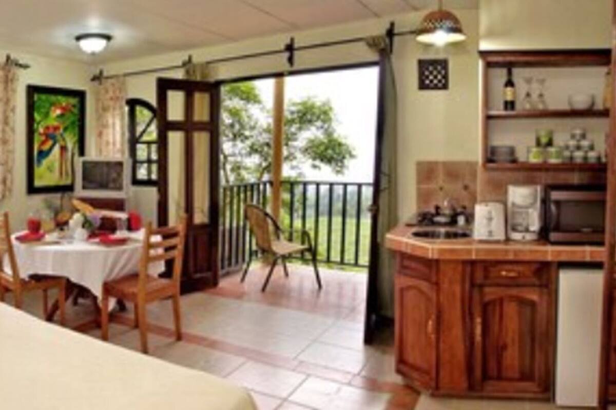 A room with two beds, a dining table with chairs, a kitchen area, and a balcony with a railing.