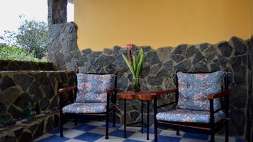 Dos sillones con estampado, un jarrón de flores sobre un piso a cuadros, frente a un muro de piedra y una pared amarilla.