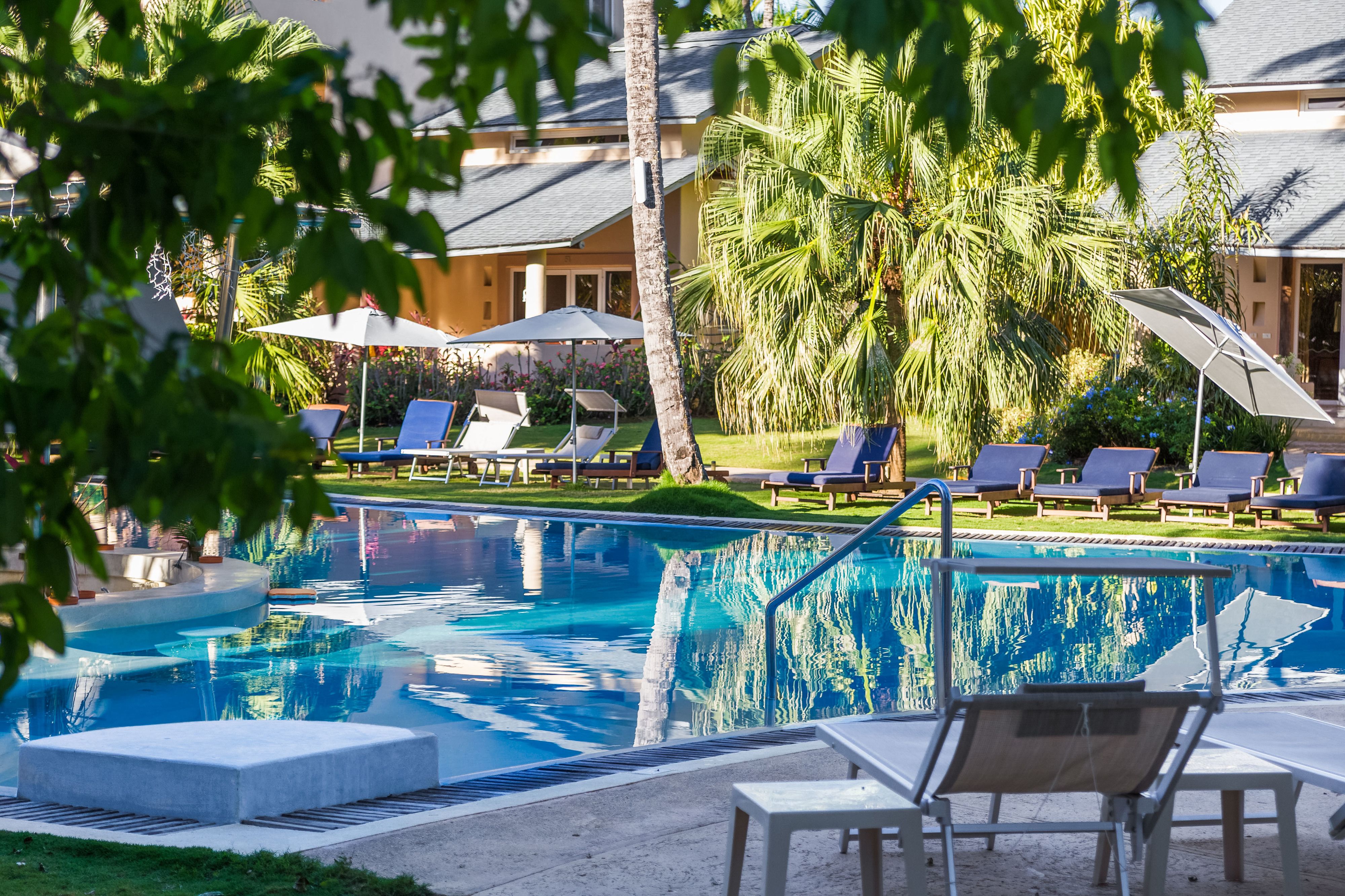 A swimming pool with lounge chairs and umbrellas, surrounded by palm trees and a building with a sloped roof.
