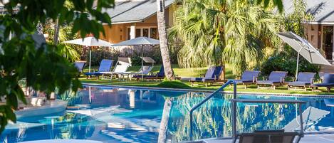 A swimming pool with lounge chairs and umbrellas, surrounded by palm trees and a building with a sloped roof.