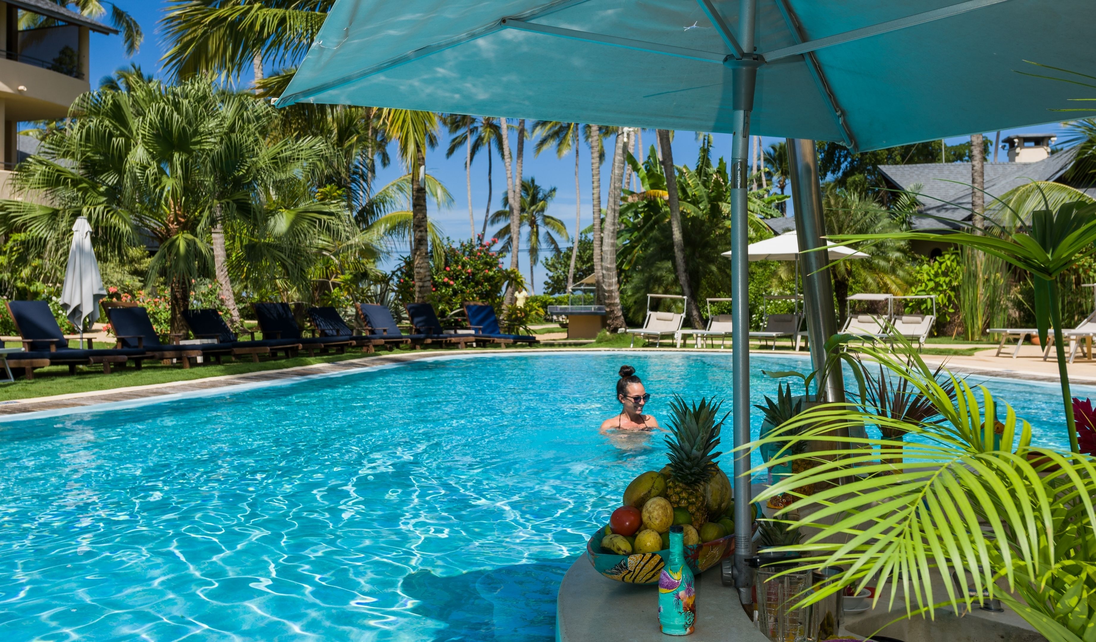 A pool area with lounge chairs and umbrellas, surrounded by palm trees and a clear sky.