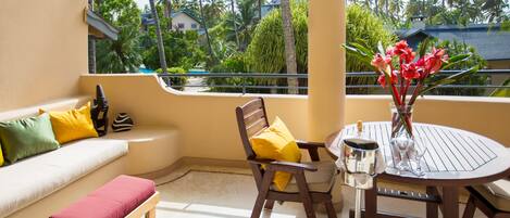 A balcony with a wooden bench, a small table with a vase of flowers, and a view of palm trees and a building.