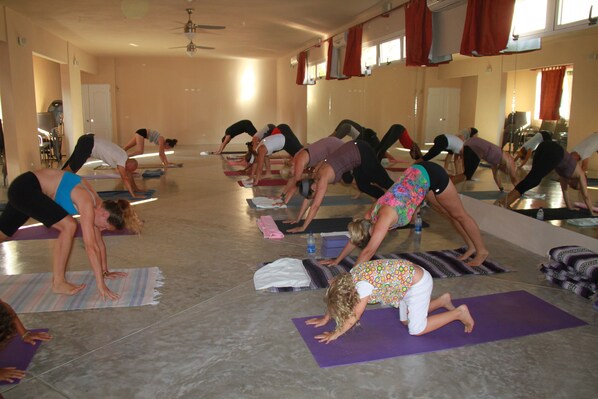 Una clase de yoga en una sala amplia con ventanas grandes, un ventilador de techo y una pared espejada.