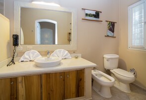 Un baño con lavamanos, espejo, inodoro, bidé, armarios de madera y una pequeña foto enmarcada en la pared.