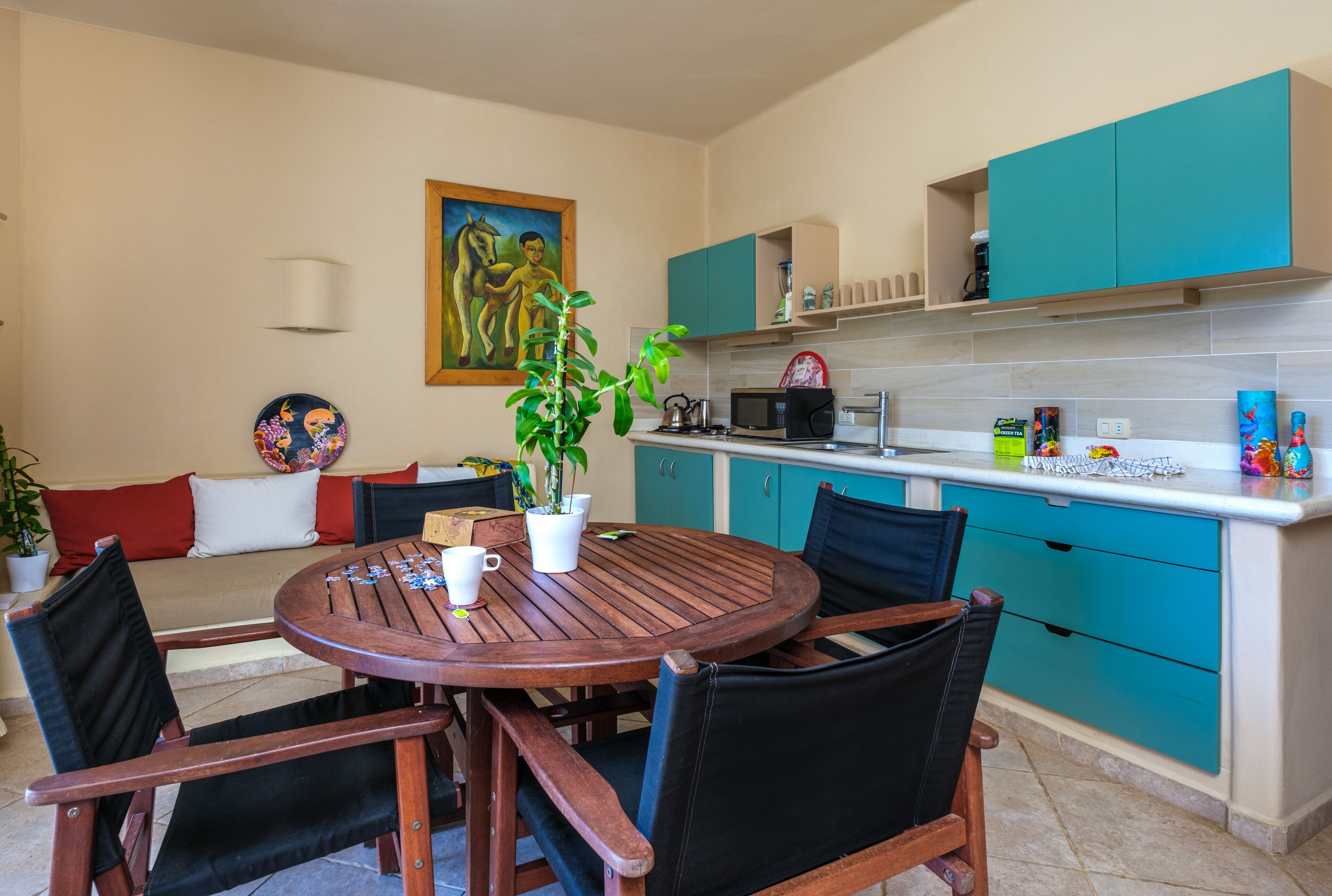Una cocina compacta con armarios color turquesa, una mesa de comedor de madera y un cuadro en la pared.