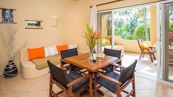 Una sala de estar moderna con una mesa de comedor de madera, sillas negras y un sofá color beige con cojines naranja. Hay un ventanal con vistas a palmeras y un balcón.