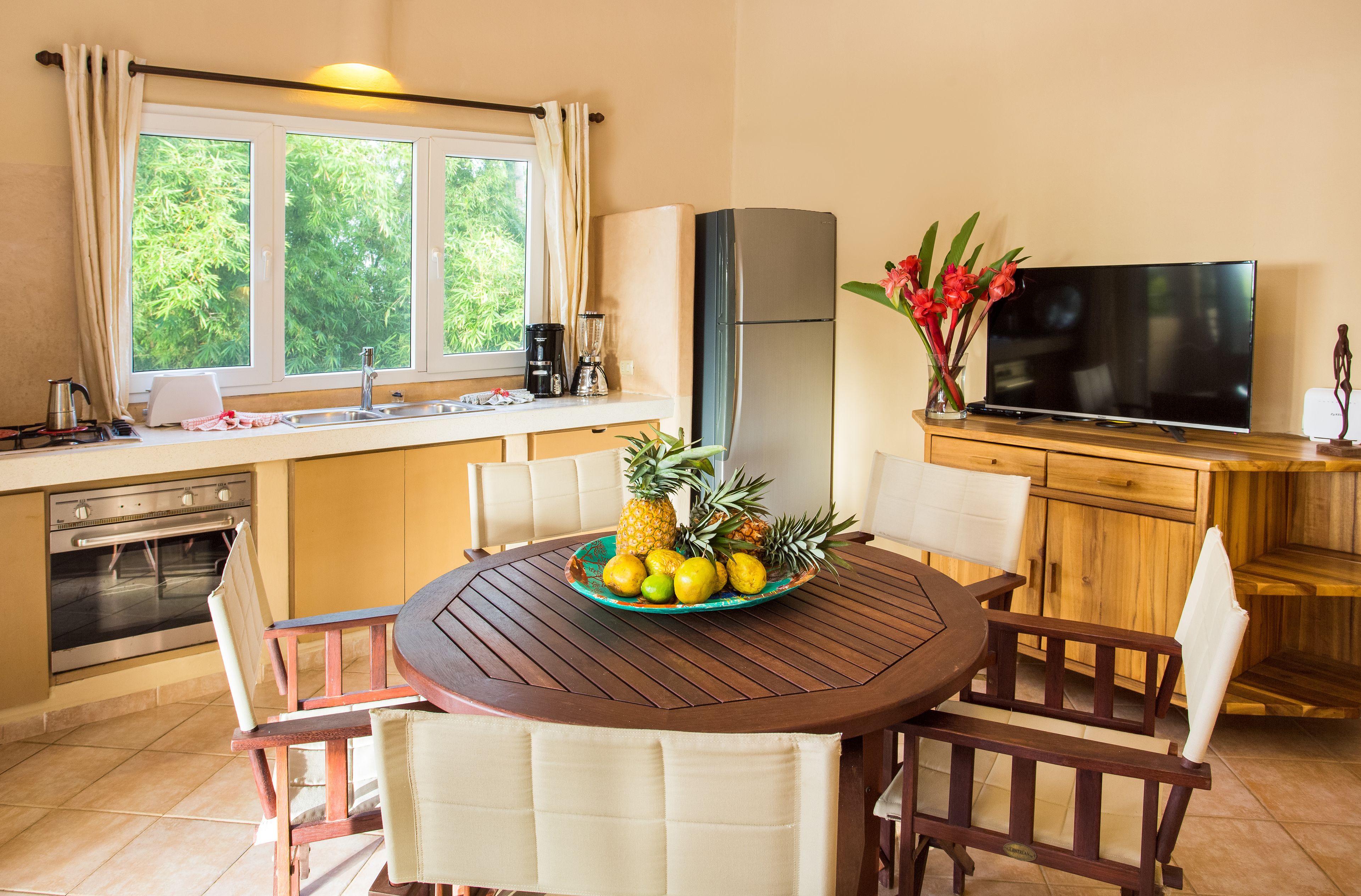 A kitchen with a dining table, chairs, a refrigerator, a TV, and a window with curtains.