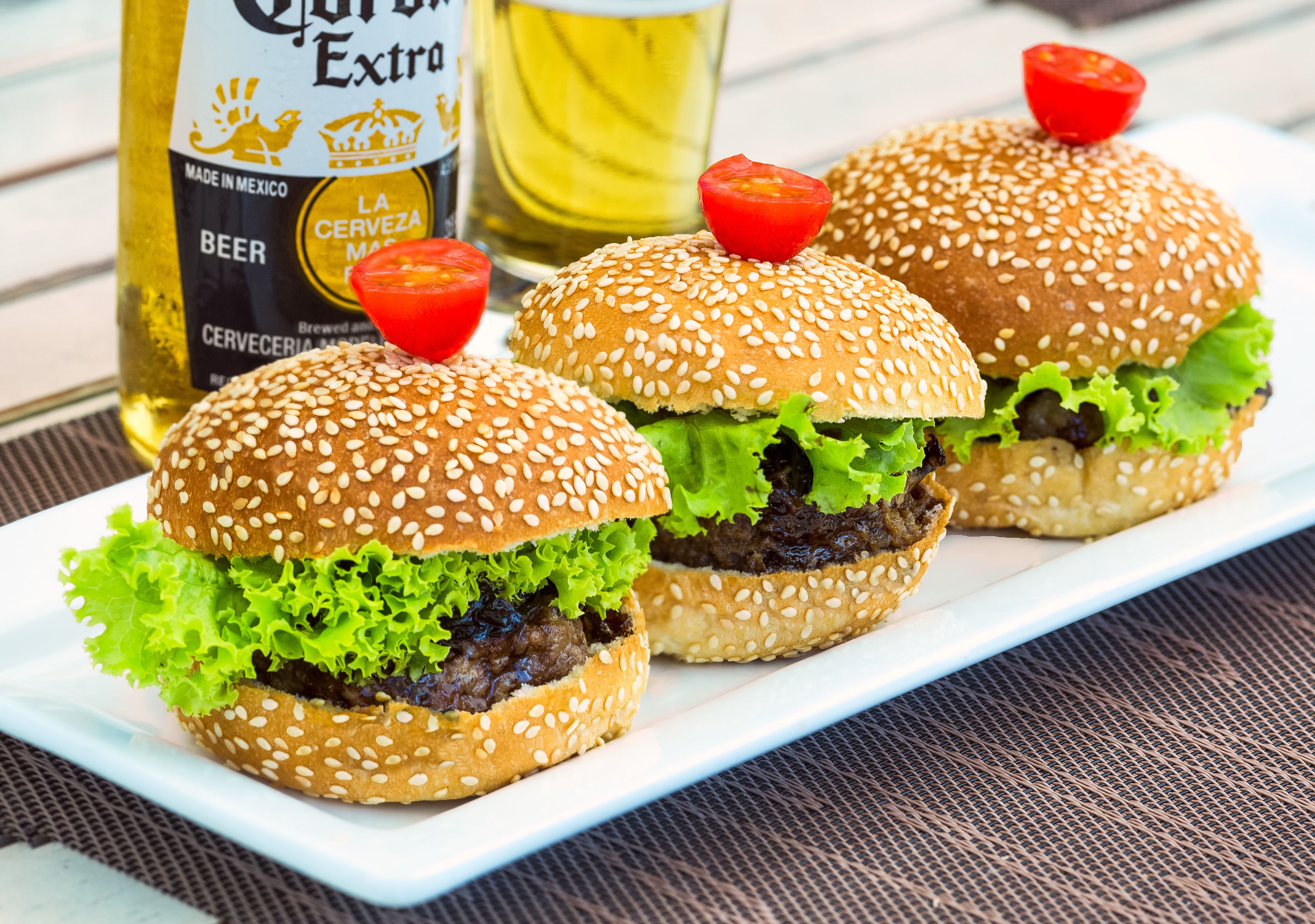 Three burgers on a white plate with sesame seed buns, lettuce, and a beef patty, accompanied by a bottle of beer and a glass of beer in the background.