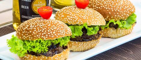 Three burgers on a white plate with sesame seed buns, lettuce, and a beef patty, accompanied by a bottle of beer and a glass of beer in the background.