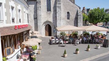 An outdoor cafe with tables and chairs, a building with a sign, and a tree in the background.
