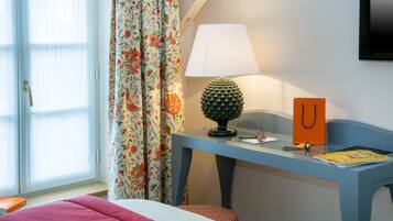 A room with a bed, a desk with a lamp, a chair, a window with curtains, and a wall-mounted TV.