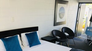 A hotel room with a bed, a desk, two chairs, a framed picture on the wall, and a door leading outside. - Blue Haven Motel (Tauranga)