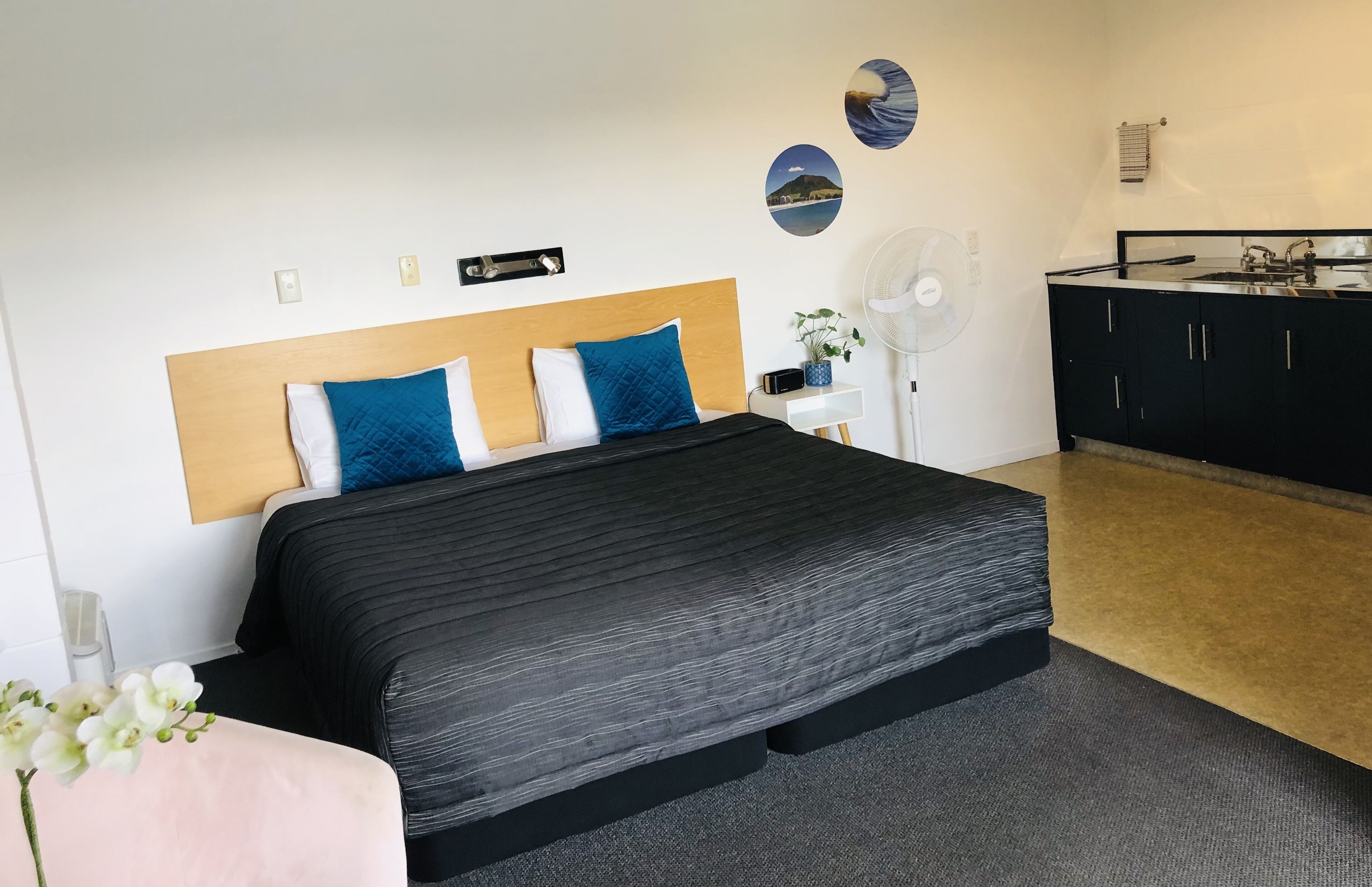 A bedroom with a bed, bedside tables, a desk, a fan, and a kitchenette.