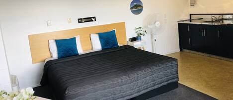 A bedroom with a bed, bedside tables, a desk, a fan, and a kitchenette.